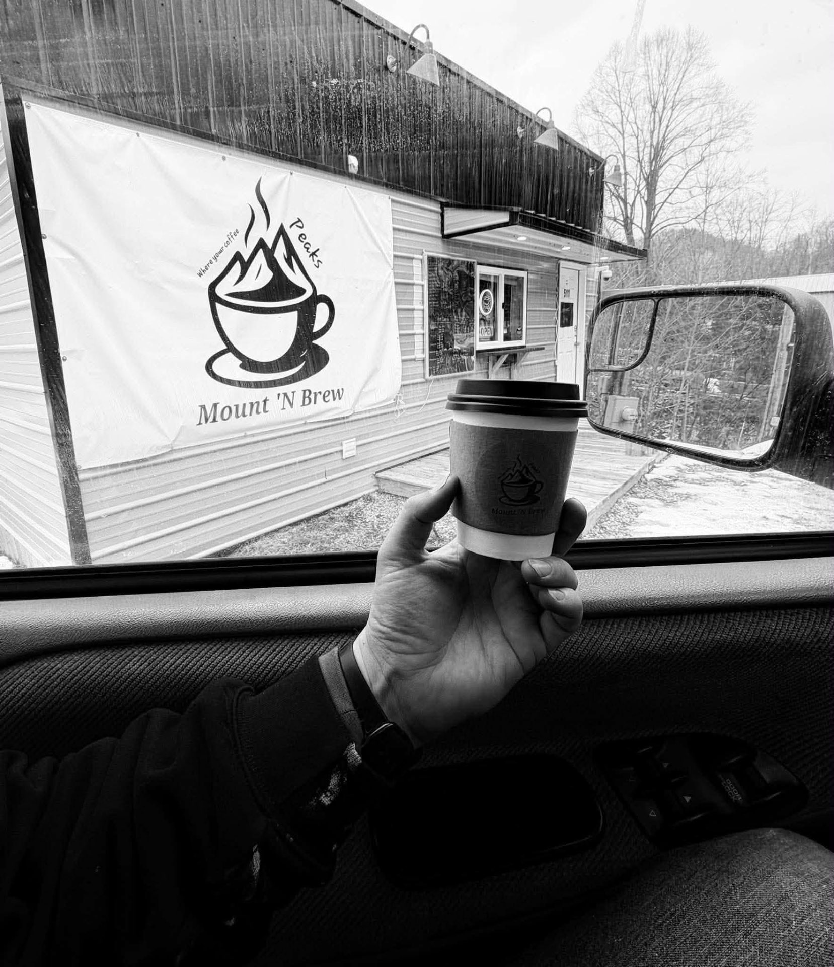 A Mount 'N Brew paper cup held up by the shop's Whitesburg storefront in the rain.